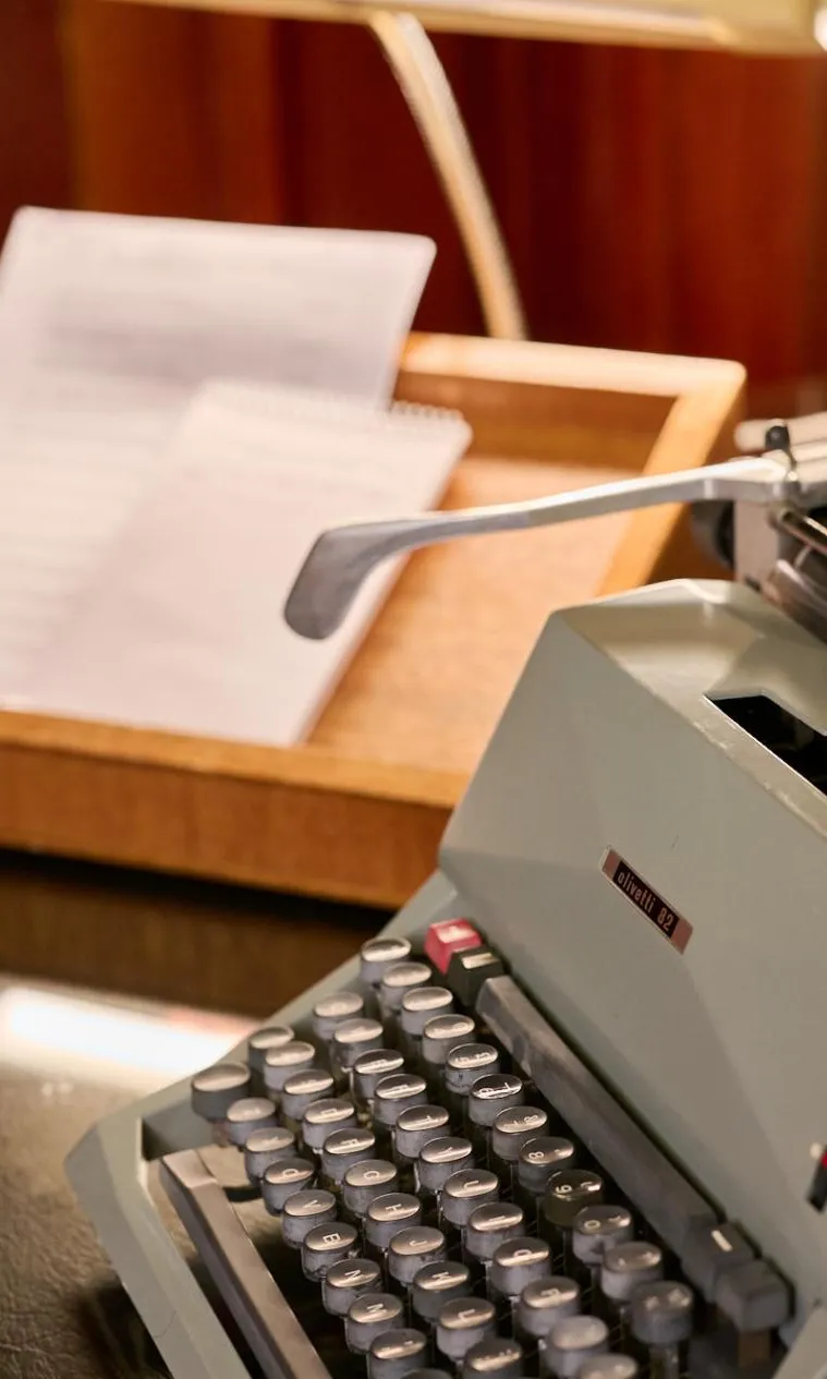 An old-fashioned grey typewriter sits on a desk next to a wooden try with documents in it. An old-fashioned grey typewriter sits on a desk next to a wooden try with documents in it.