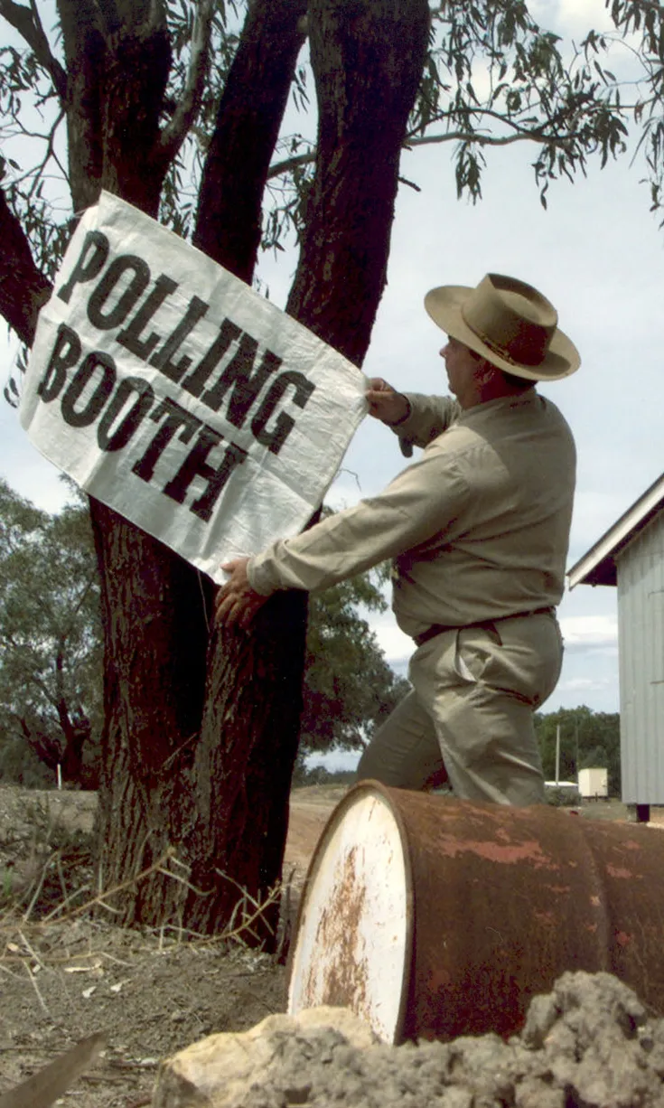 A man in a hat puts a 'Polling Booth' sign up on a gum tree which is next to a corrugated iron shed and water tank on an outback property.