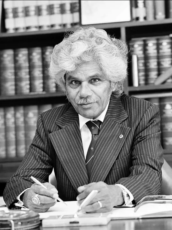 Neville Bonner sits at his desk, wearing a striped suit and tie, a pen in his hand, behind him a full bookshelf.