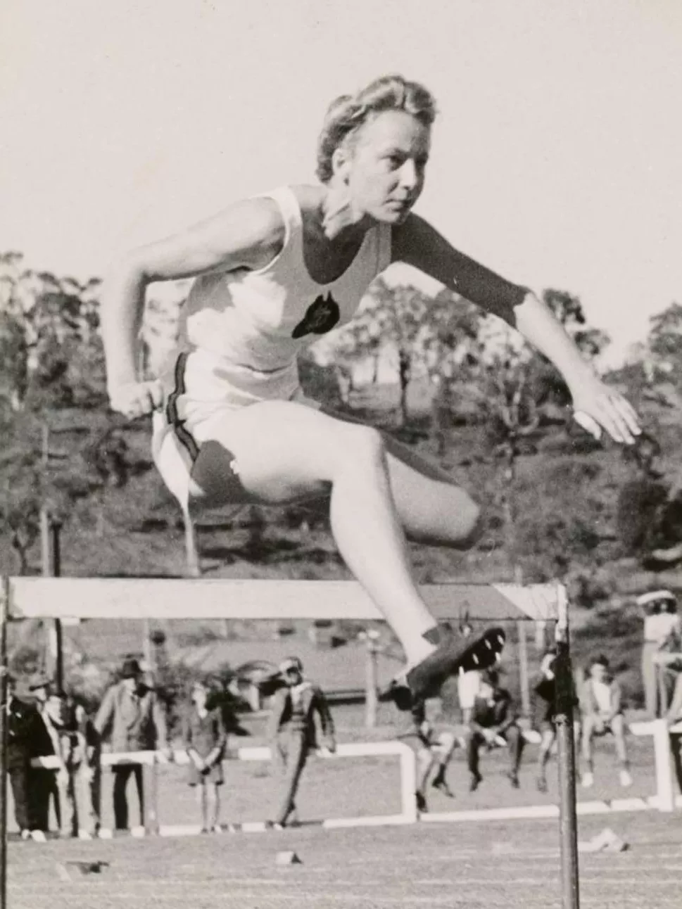 A young woman wearing a white singlet and shorts, jumps over a hurdle on a race track.