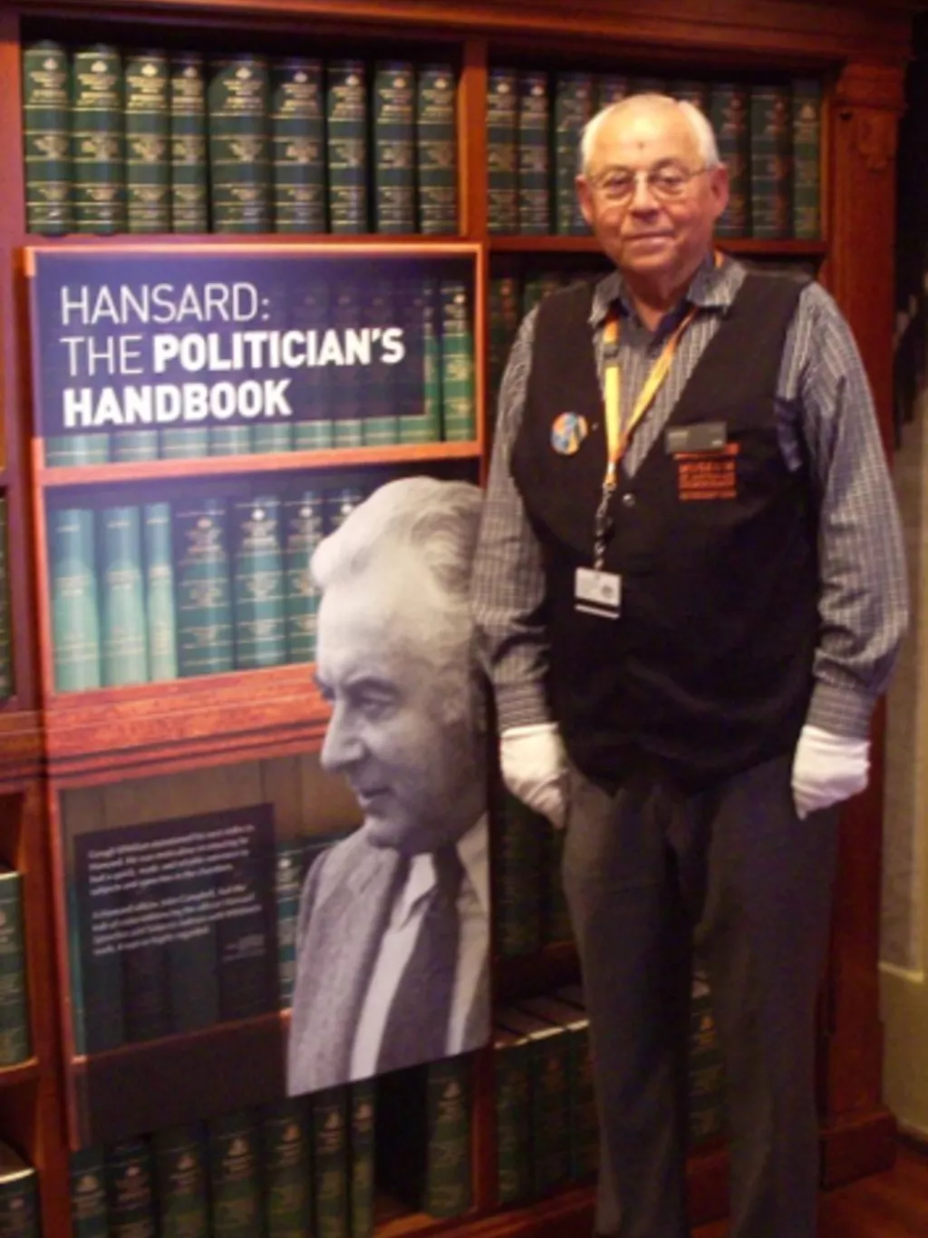 John Campbell as a MoAD volunteer guide, standing next to shelves full of bound hansard volumes.