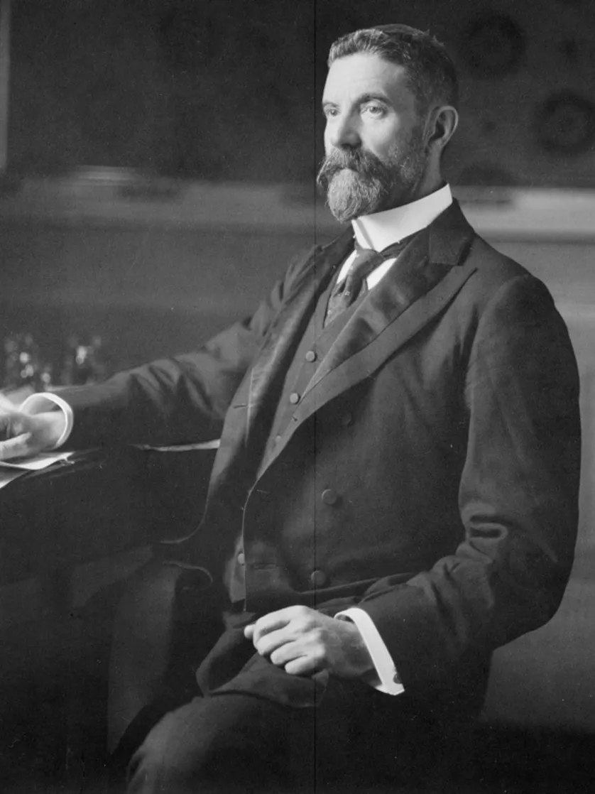 Alfred Deakin, wearing a three-piece suit and seated at a desk covered in paper documents.