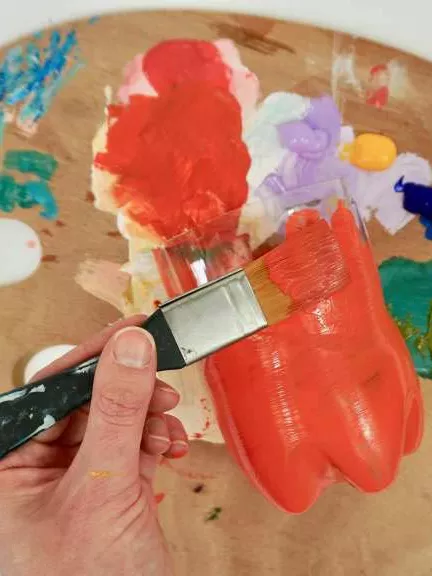 A close up of a hand holding a paint brush painting a plastic bottle red.