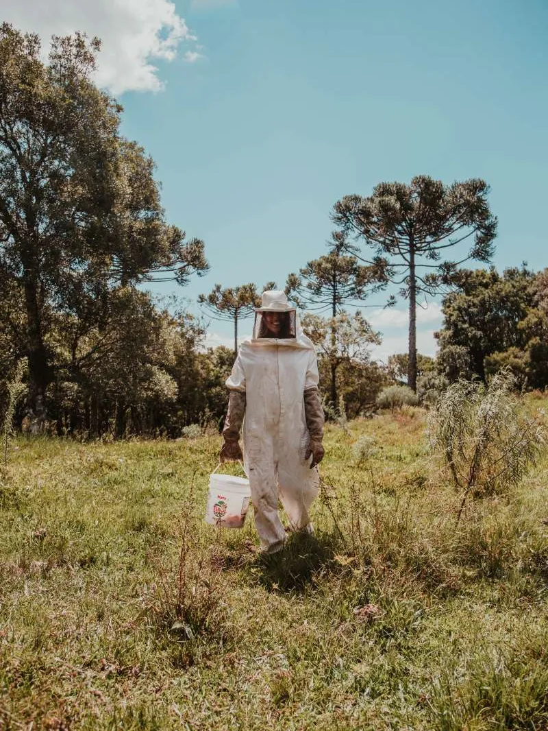 A person standing in a field wearing a beekeeper suit and holding a bucket.