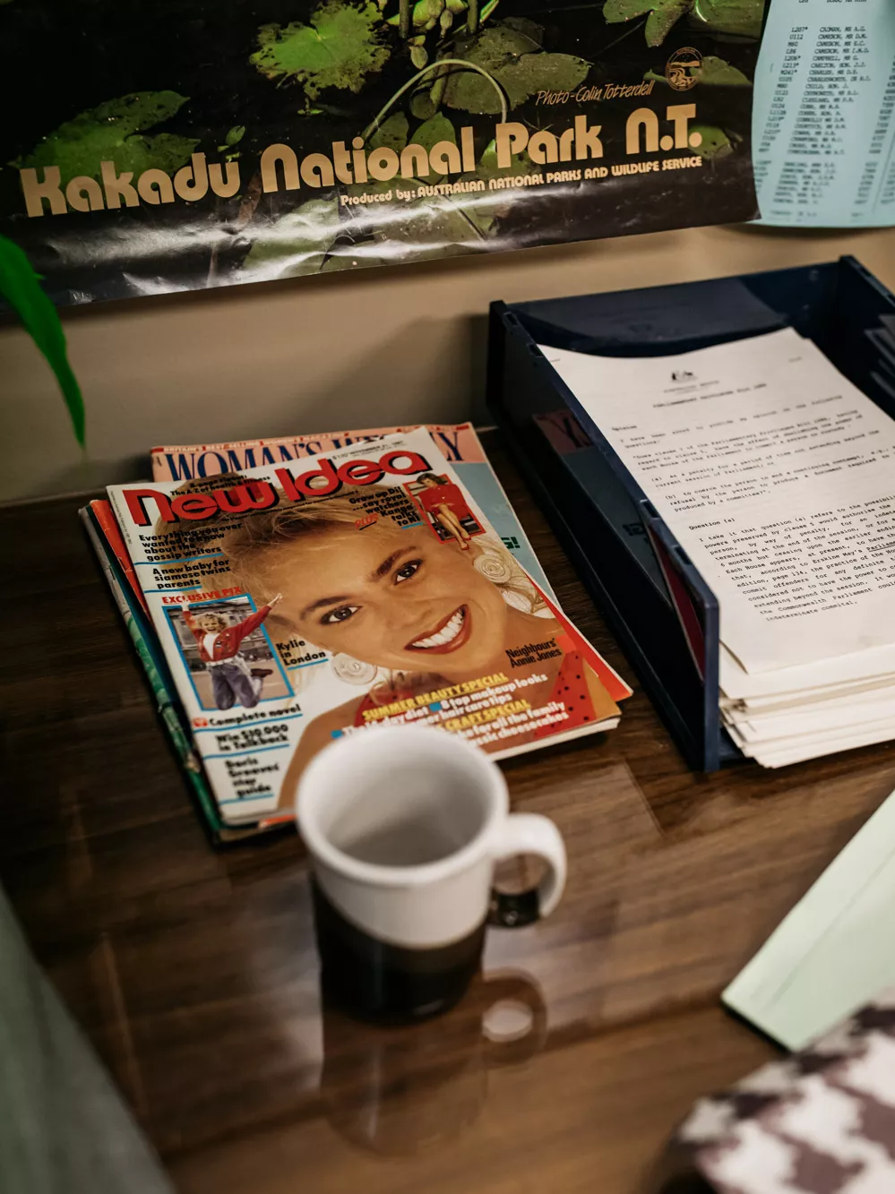 A staco of magazines and a coffee cup sat on one of the desks, alongside a tray of papers. On the wall behind, the bottom of a 'Kakadu National Park' poster can be seen.