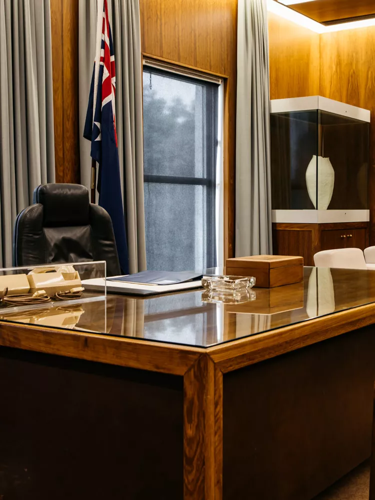 A colour photograph of the Prime Minister's Desk. Behind it is a comfortable office chair and an Australian flag on a pole. In front and to the side of the desk are armchairs.