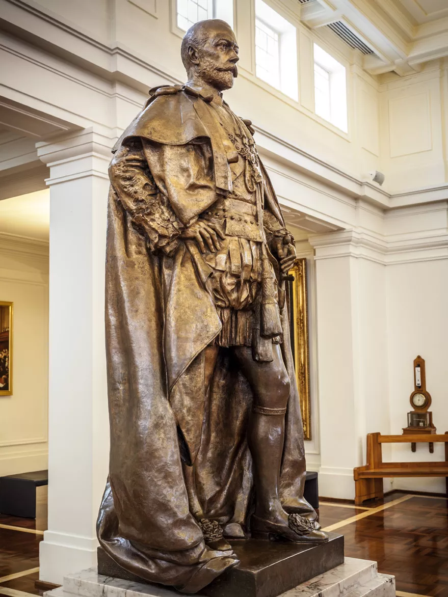 A bronze statue of a man with a cloak on a marble pillar in a room with timber floors.