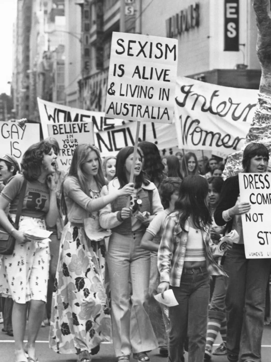 A black and white photo of a group of women holding protest signs in 1975. Signs say 'sexism is alive and well in Australia' and 'dress for comfort not style'.