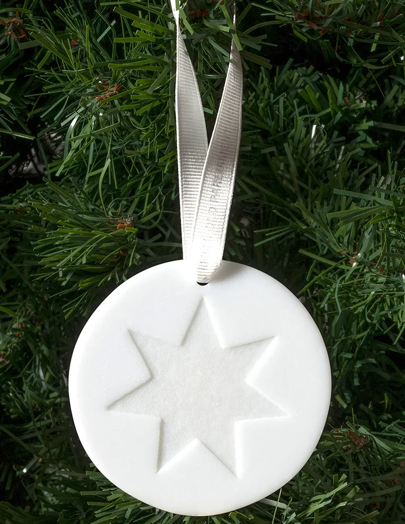 A circular white ornament with a six-pointed start in the centre, hanging by a ribbon against plastic pine leaves. 