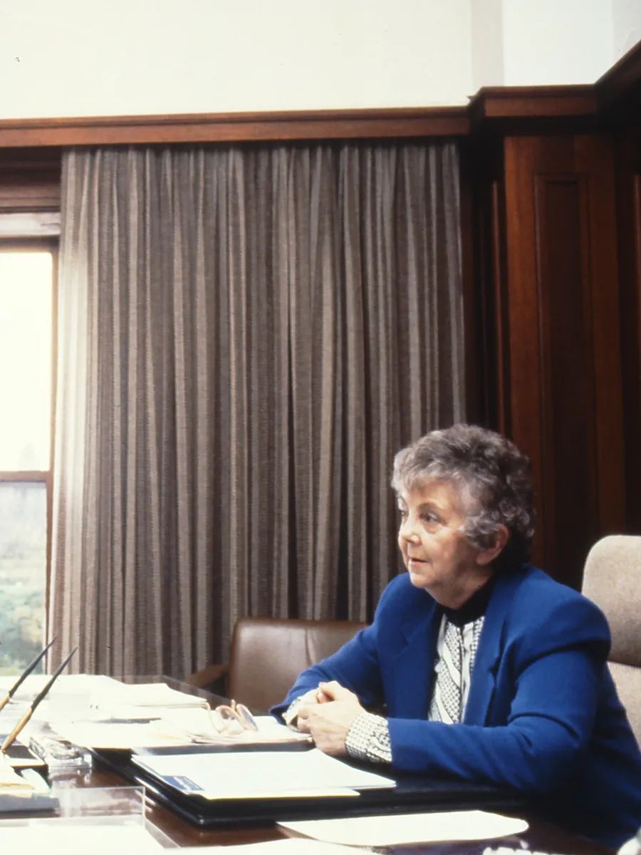 This professional colour photograph shows Principal Private Secretary John Porter and Speaker Joan Child sitting across from each other at the Speaker’s desk. John is wearing a dark suit, white shirt and dark tie and Joan is in a white blouse and vibrant blue jacket. They are discussing the daily program for the House of Representatives Chamber. There is a vase of roses, stationery and paperwork on the desk. In the background the curtains have been drawn to reveal a large east-facing window.