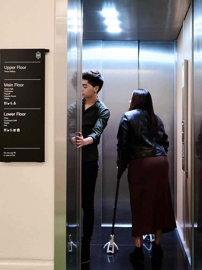 A man holds the door for a woman on crutches inside a lift at Old Parliament House, there is a sign outside the lift for the different floors of the building.