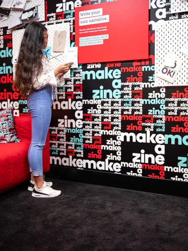 Kids stand in front of a wall with black, white, red and blue wallpaper with the words 'make a zine'. 
