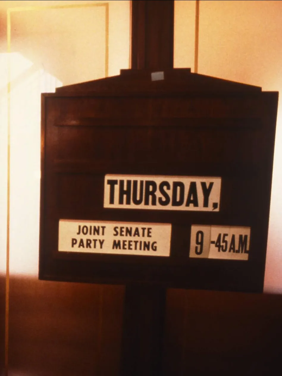 A wooden sign with the text 'Thursday, joint Senate party meeting, 9.45 a.m.' stands outside the door of a room with red carpet in Old Parliament House.