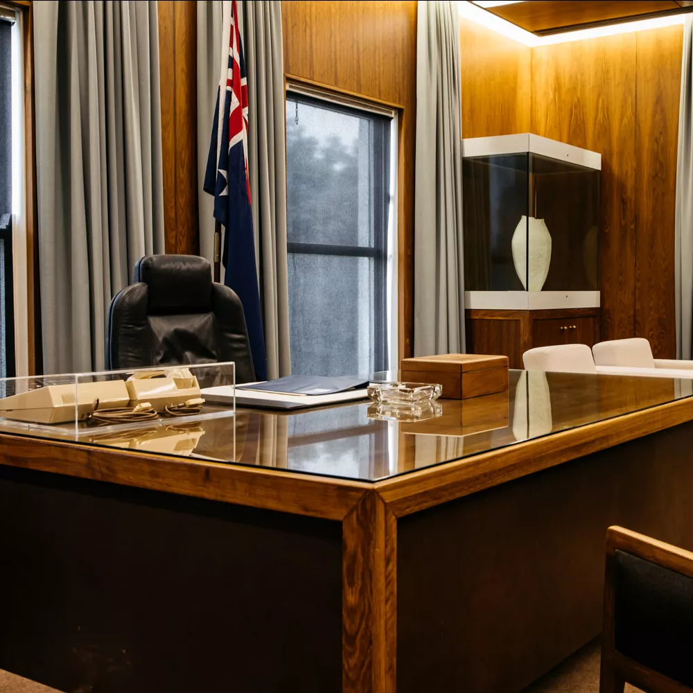 A black bean wooden desk with a glass cover in a room with an Australian flag, leather chair and grey curtins.