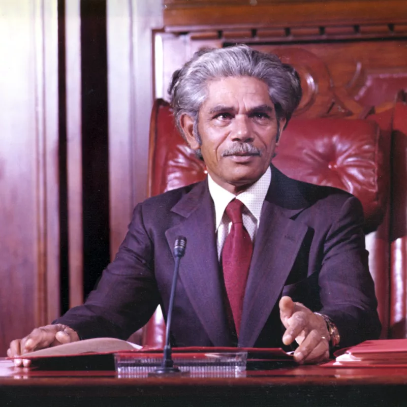 Nevill Bonner seated in the Senate chamber, wearing a suit and red tie.
