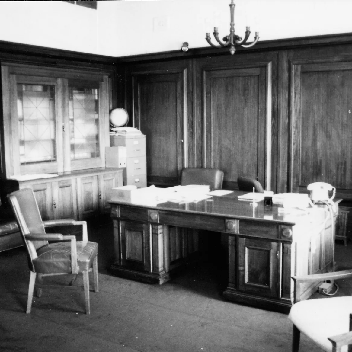 This black and white photograph shows the layout of the Clerk of the Senate’s office in 1985 when it was occupied by Alan Cumming Thom. The timber-panelled room contains a large desk (originally designed by the building’s architect, John Smith Murdoch), glass-fronted built-in bookshelves and a sideboard with an indoor plant. There are a range of visitor chairs and an easy chair in the foreground.