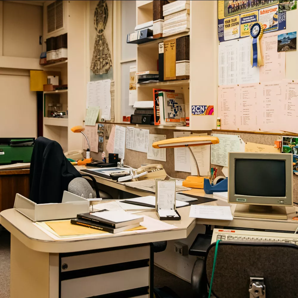 Two desks cluttered with books, pens, staplers, old fashioned 80s computers, and a pinboard covered in papers. 