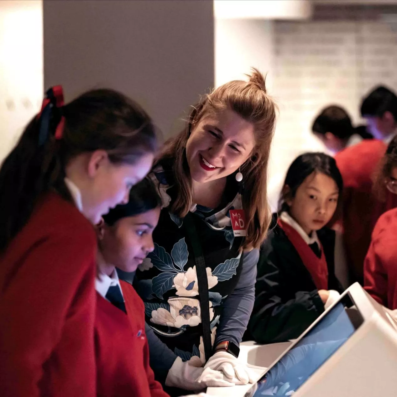 High school students in red jumpers look at screen in front of them and a MoAD educator stands next to them.