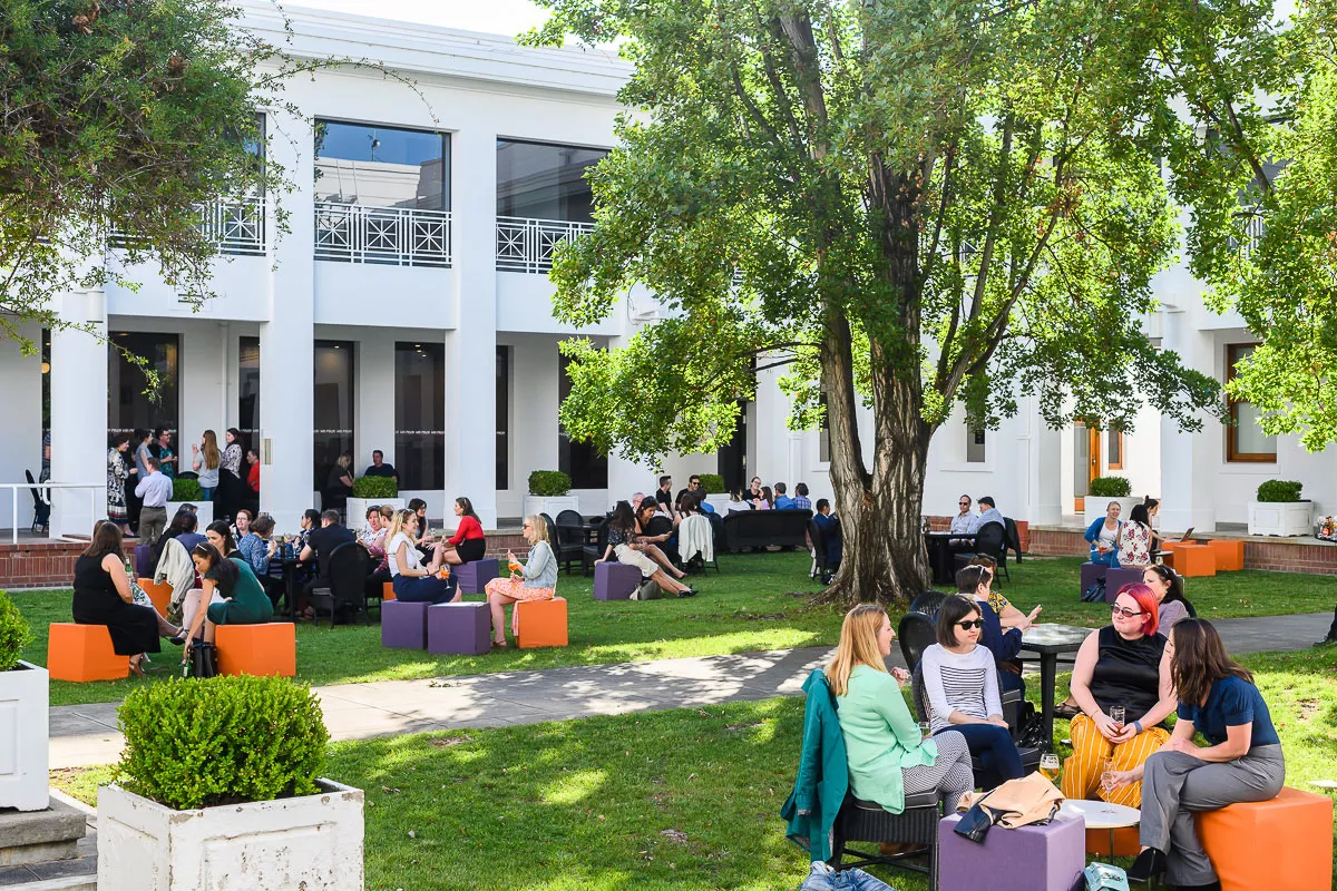 A green grassy courtyard surrounded by white walls, with trees and people sitting at tables. 