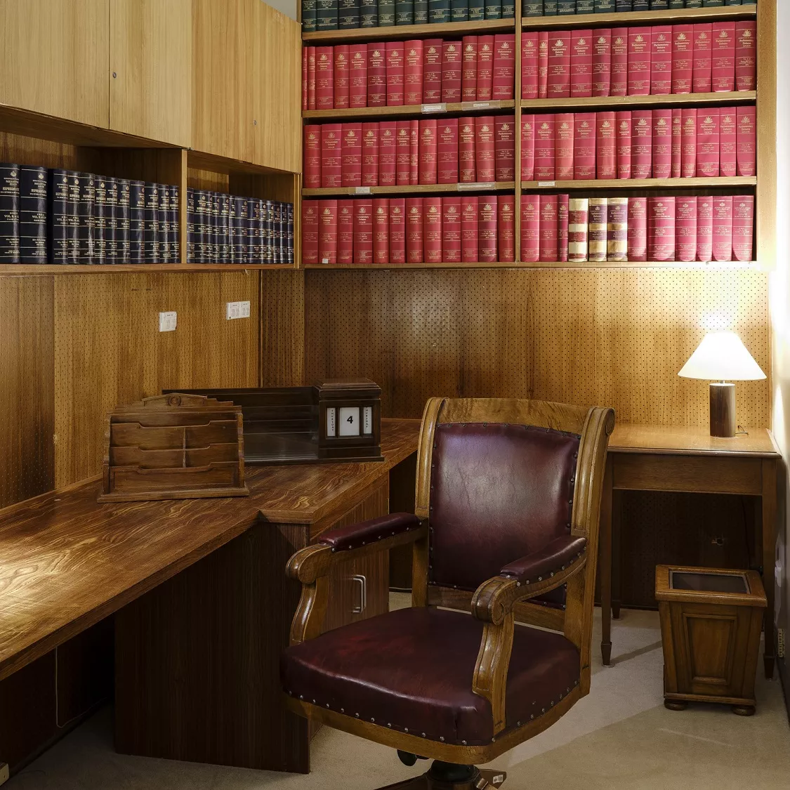 A leather-cushioned wooden swivel chair, next to a long wooden desk with a lamp and desk calendar; on two walls there are inbuilt bookcases with rows of bound books.