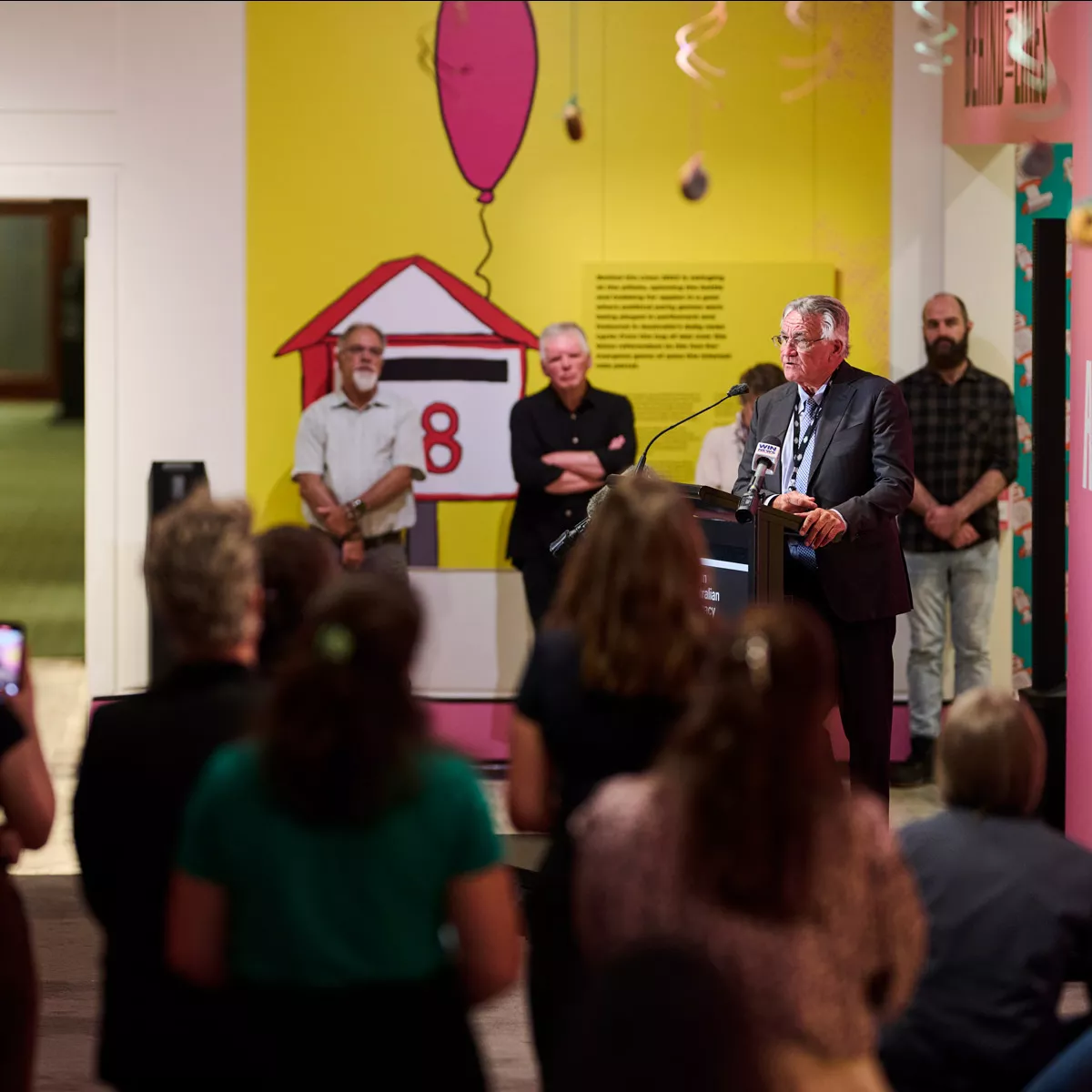 Barrie Cassidy stands behind a podium and speaks to an audience, some of whom are recording on photos and cameras. 