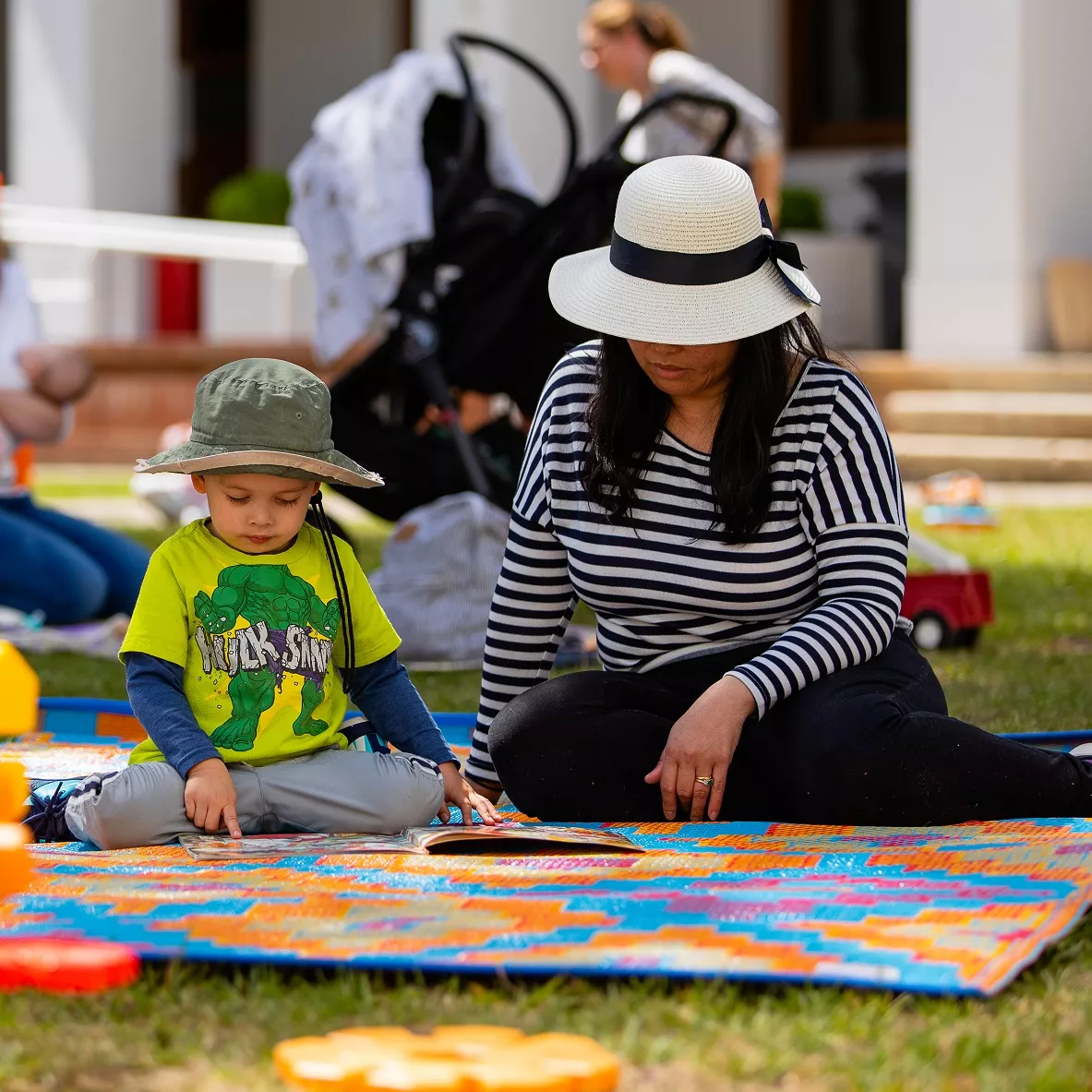 A child reading a book with their adult carer on a colourful mat on the lawn.