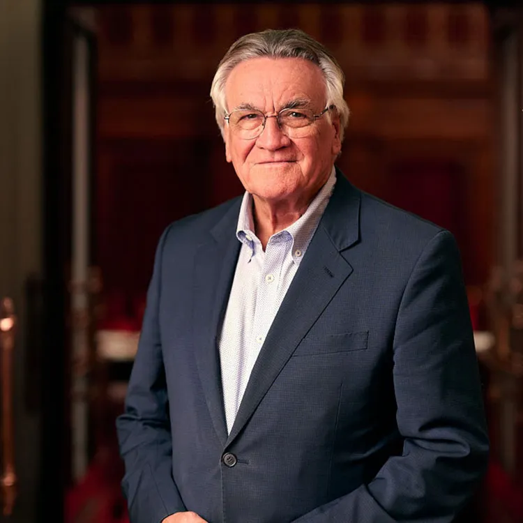 Barrie Cassidy wears a blue suit with light blue shirt. He is standing in Old Parliament House in an ornate wooden doorway.