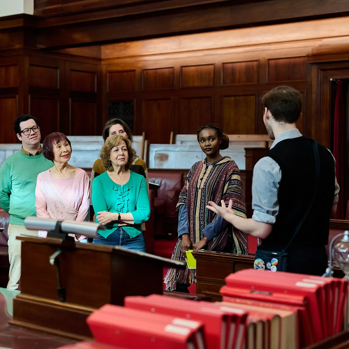 A tour guide faces a group of people who listen to what he is saying. They are standing in front of the red leather bench chairs of the Senate Chamber.