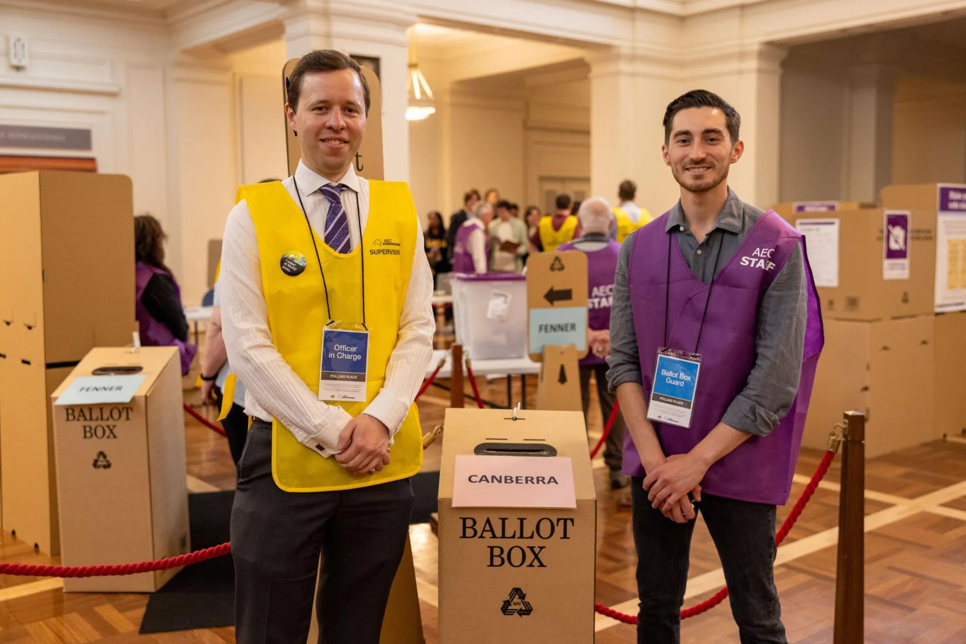 Two AEC officials, one in a yellow vest and one in a purple vest, stand beside a cardboard ballot box for Canberra in King's Hall in Old Parliament House.