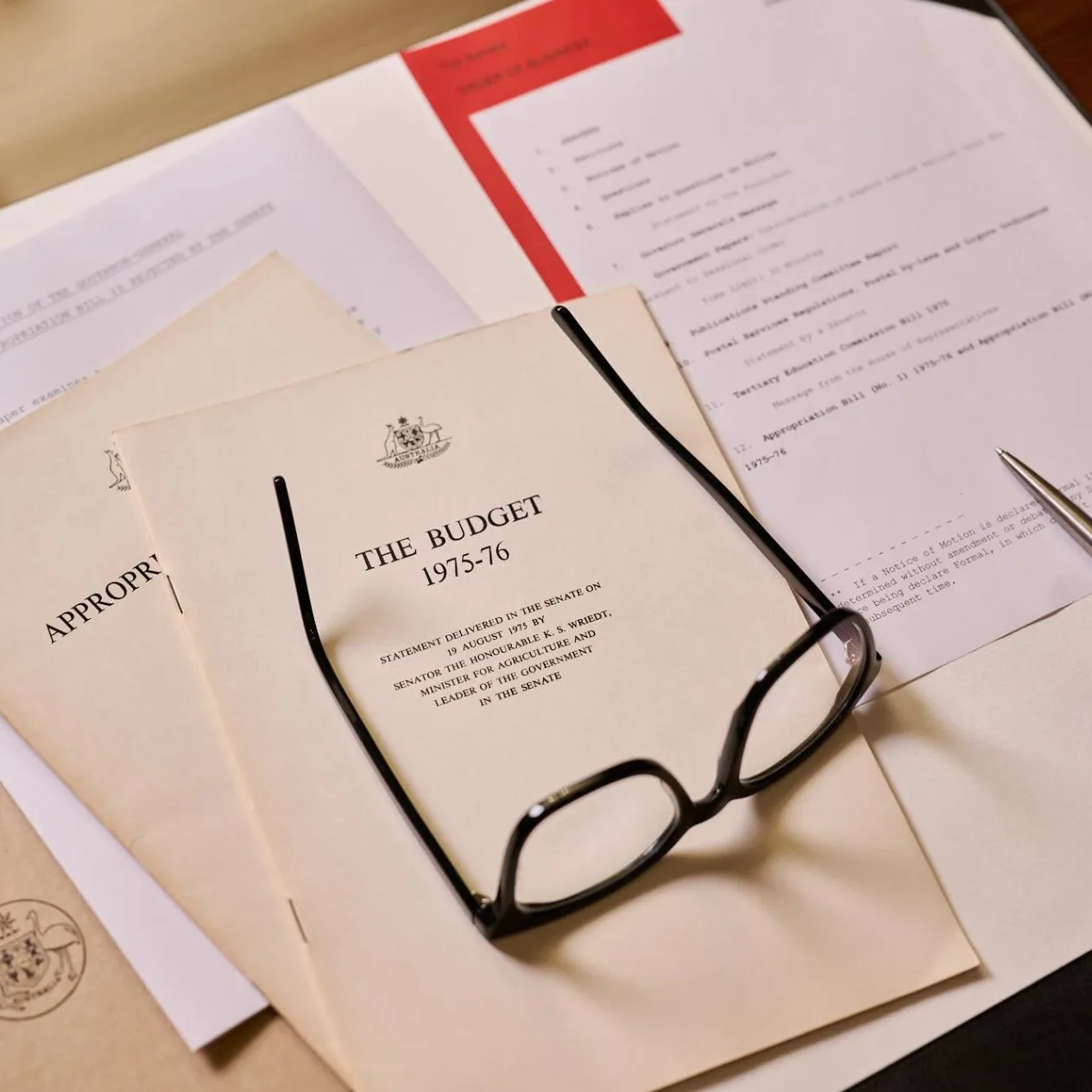 A stack of papers on a desk, a yellow booklet titled 'The Budget 1975-76' with the Commonwealth Coat of Arms is on top, along with a pair of black-framed reading glasses and a pen.