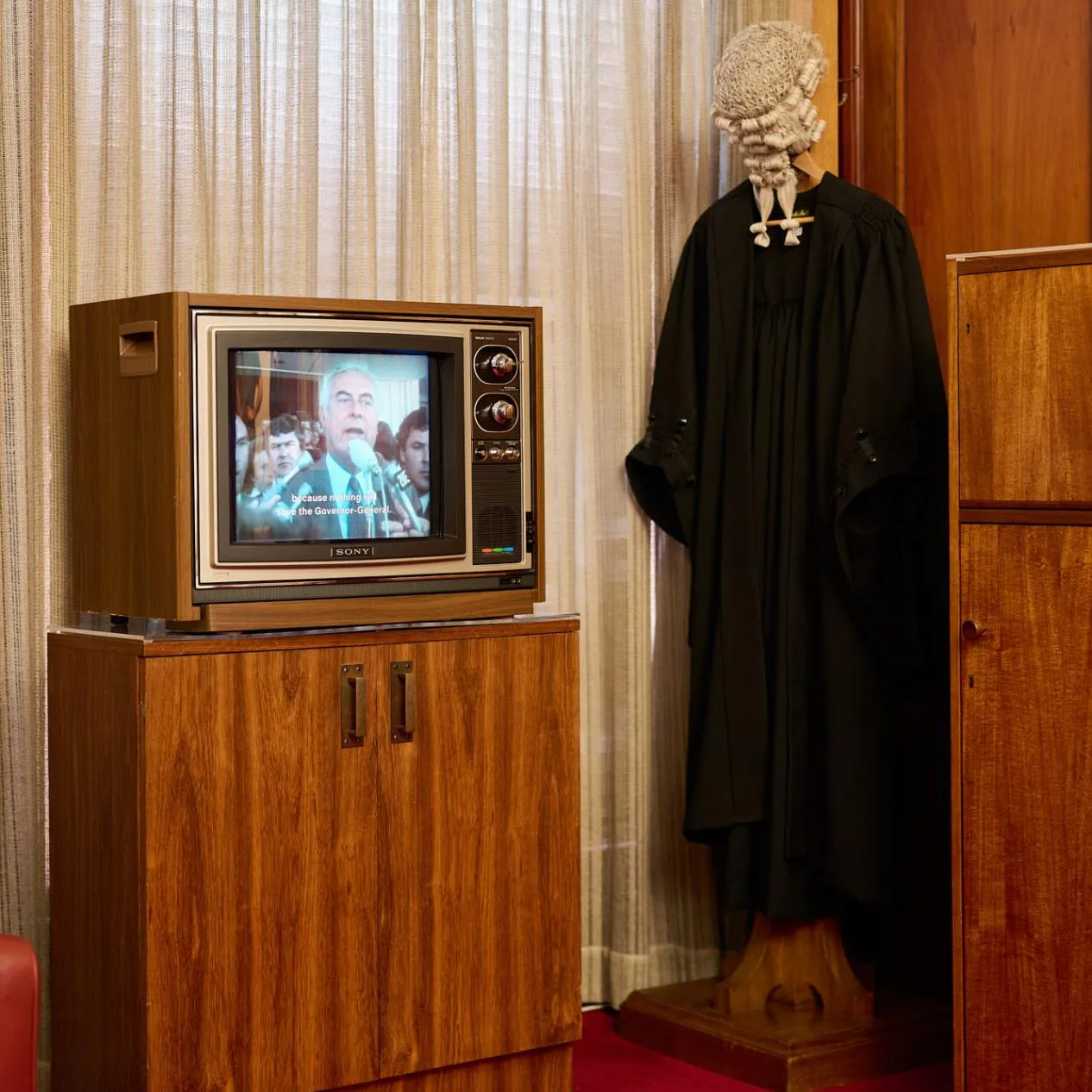 In a room in Old Parliament House a wooden TV with an image of Gough Whitlam is on a wooden cabinet next to a stand with a black gown and white wig. There is red carpet on the floor.