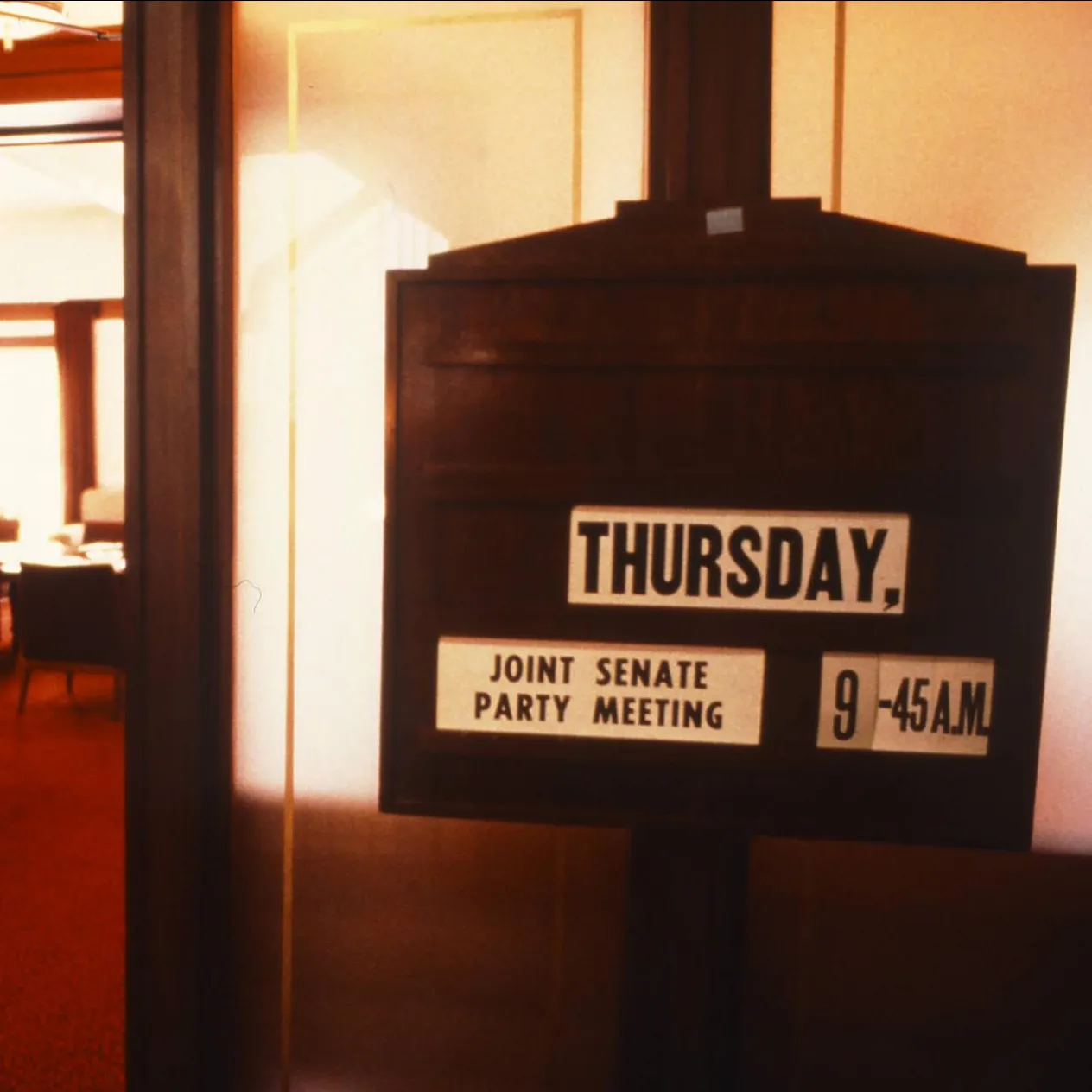 A wooden sign with the text 'Thursday, joint Senate party meeting, 9.45 a.m.' stands outside the door of a room with red carpet in Old Parliament House.