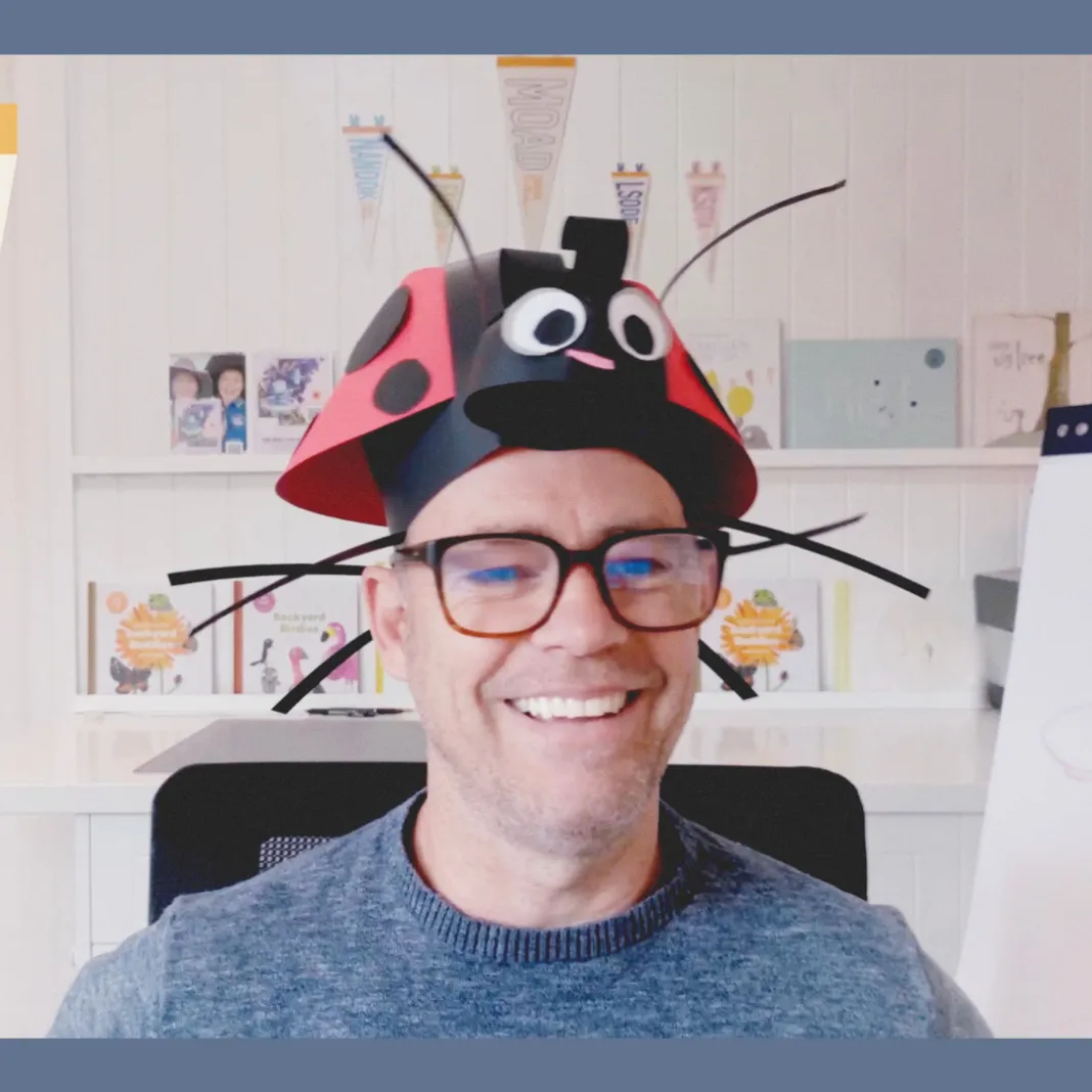 Andy Geppert in front some bookshelves with his books on them, with a lady bug made of paper on his head.