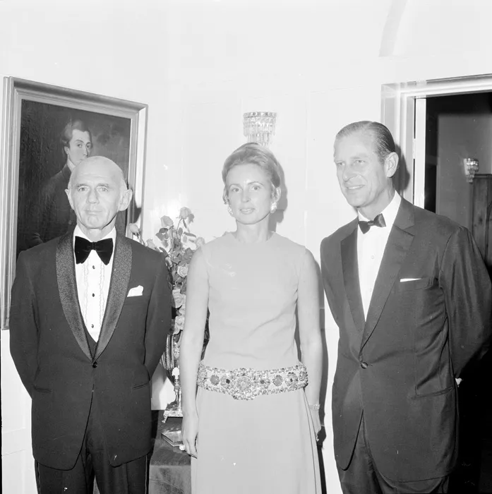 This black and white group photograph captures Prime Minister McMahon in a tuxedo with ruffled white shirt and black velvet bow tie standing next to his wife Sonia who is wearing a sleeveless light-coloured dress with heavily jewelled belt. Her long blonde hair is piled up in a chignon and she is wearing large earrings. To her left is Prince Philip in a more restrained tuxedo and bow tie. The group are standing in an elegant room and there is a flower arrangement and oil painting in the background.  