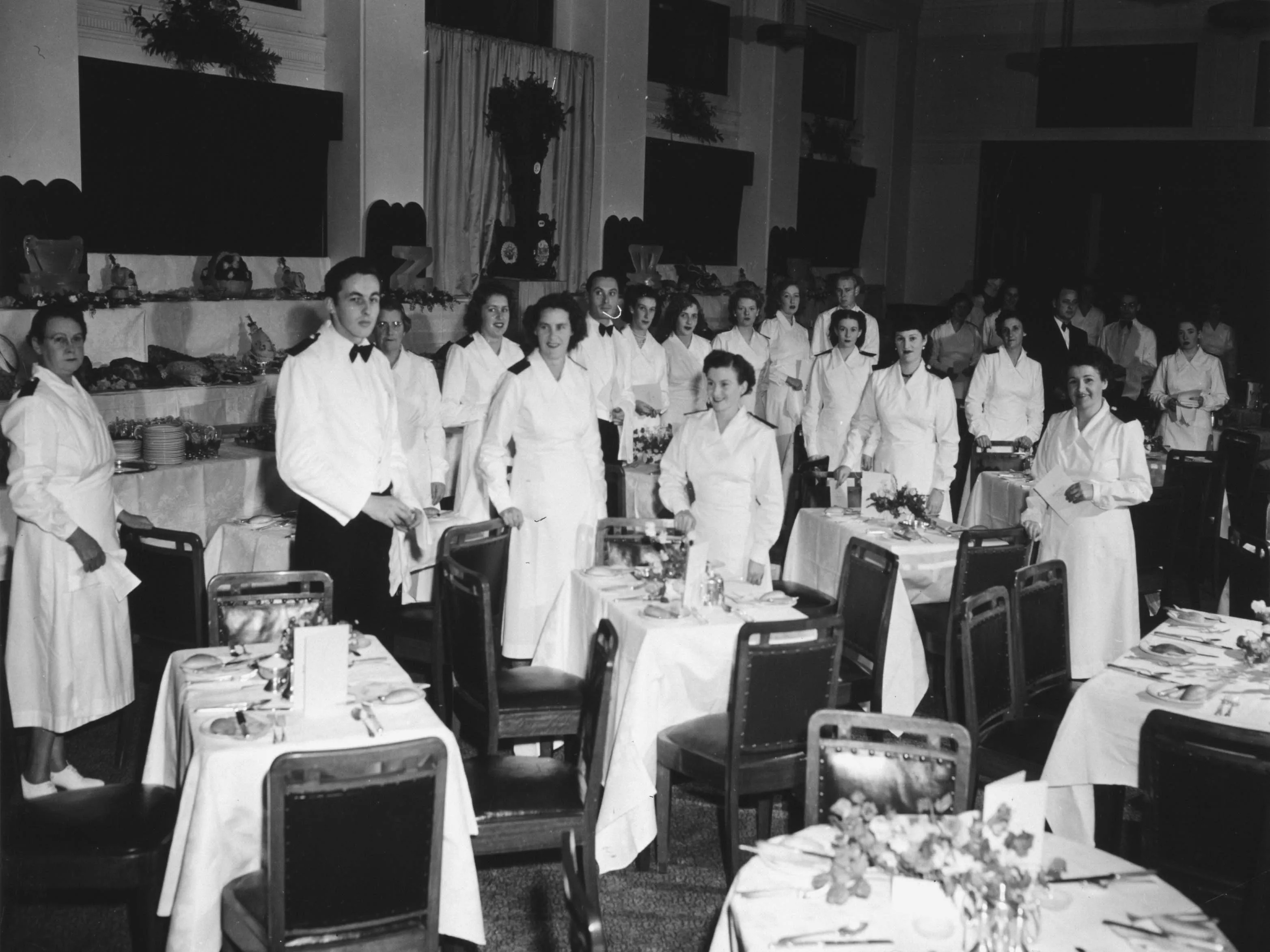 This black and white photograph captures the moment before a dinner  celebrating the 50th anniversary of Federation in the Members’ Dining Room at Parliament House. Small dining tables seating two to four guests are set up in the foreground with white linen tablecloths and napkins, silverware, crockery, bread rolls, floral arrangements and menus. In the background are tiered tables laden with food and crockery and topped with ice sculptures, centrepieces and floral arrangements. In the midground, 23 members