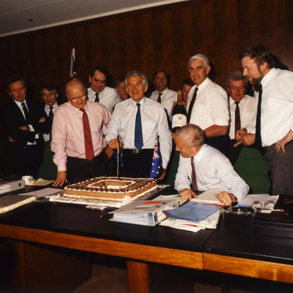 Prime Minister Bob Hawke stands and cuts a square cake, surrounded by a group of men all smiling.