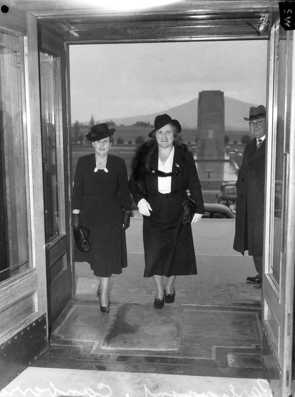 A black and white photo of Dorothy Tangney and Enid Lyons wearing dress suits and hats walking in through the front door of Old Parliament House.