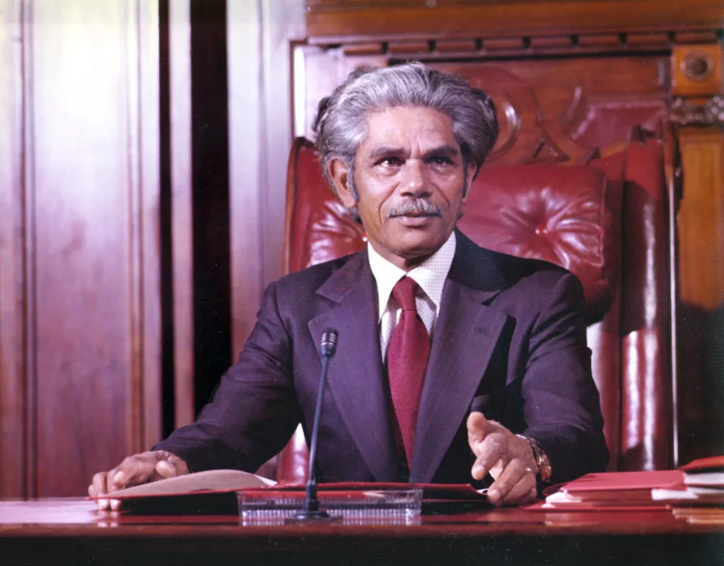 Nevill Bonner seated in the Senate chamber, wearing a suit and red tie.