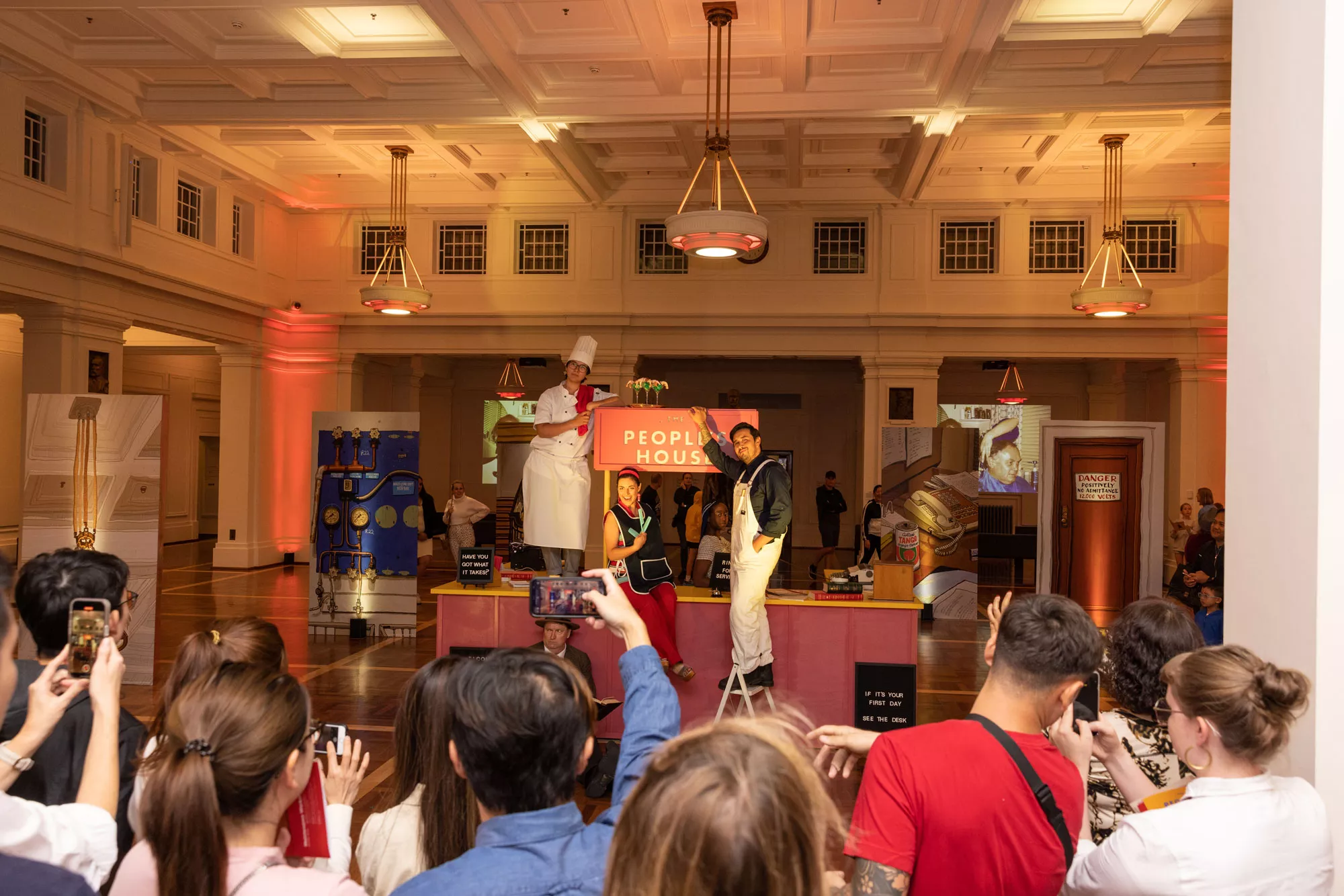 A group of visitors hold up their phones and film a performance with actors dressed as a chef, a hairdresser and a handyman inside King's Hall with a sign in the background that says The People's House. 