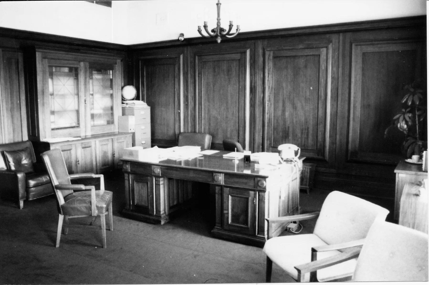 This black and white photograph shows the layout of the Clerk of the Senate’s office in 1985 when it was occupied by Alan Cumming Thom. The timber-panelled room contains a large desk (originally designed by the building’s architect, John Smith Murdoch), glass-fronted built-in bookshelves and a sideboard with an indoor plant. There are a range of visitor chairs and an easy chair in the foreground.