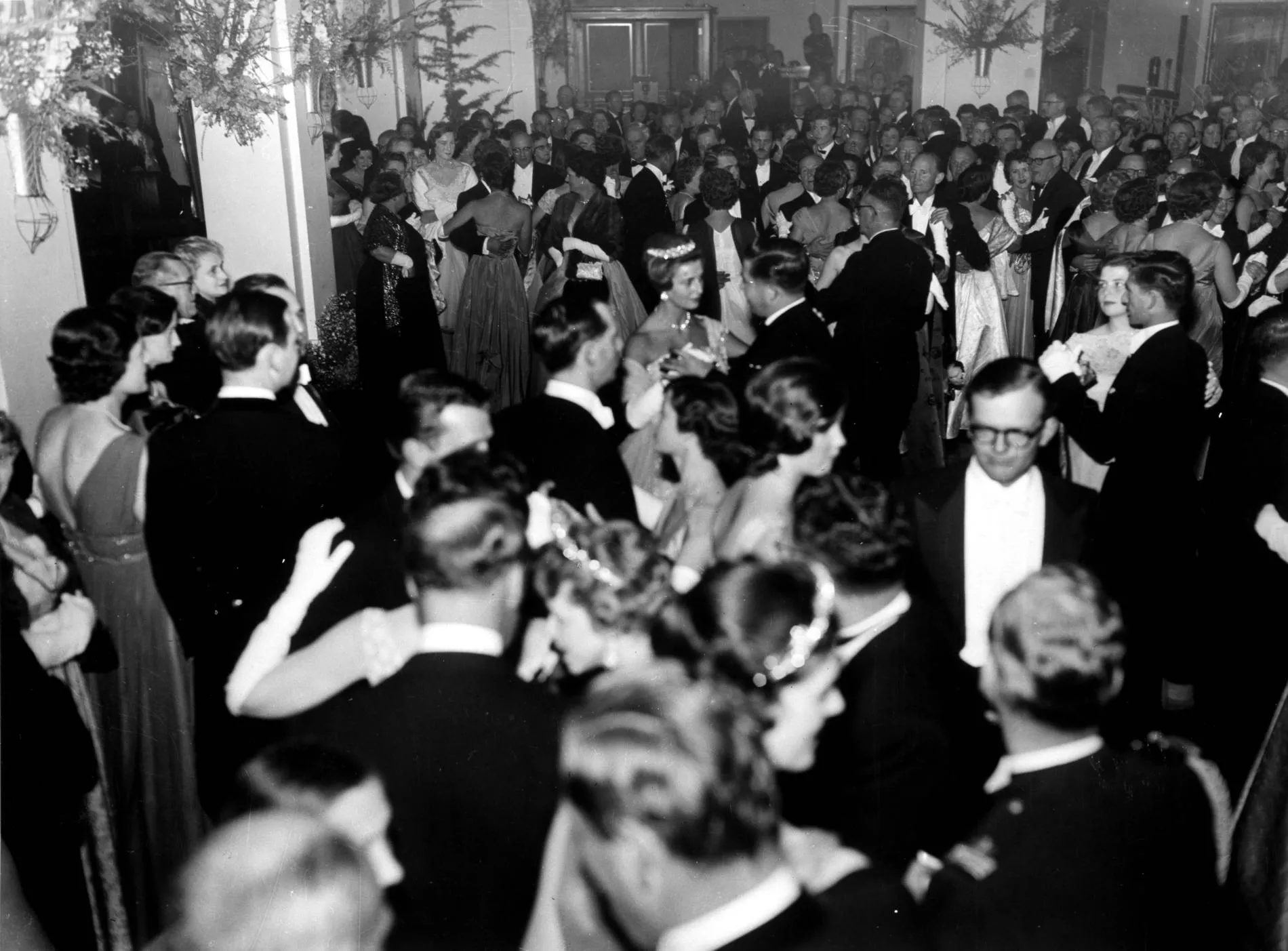 In this black and white photograph a beautifully decorated King’s Hall is packed with people. In the background and left of the frame people stand and chat on the edge of the dance floor while those in the centre of the image and on the right are dancing what appears to be a waltz. Princess Alexandra in a sparkling tiara is prominent in the centre of the crowd. The men are in tuxedos and the women wear evening dresses with elbow length white gloves.