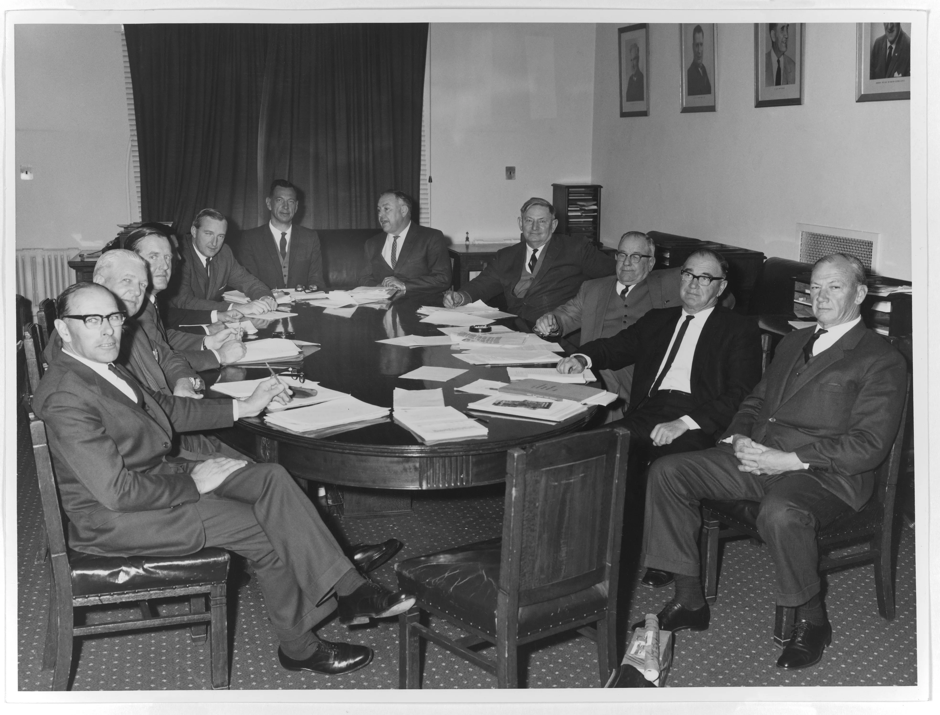 This black and white photograph shows 11 members of the Country Party Standing Committee on Drought meeting in the party room. They are seated at a large oval table covered with papers and almost all of the men have looked up to acknowledge the camera. They are uniformly dressed in dark suits, white shirts and ties and some are wearing waistcoats. In the background is a large curtained window, small desks with stationery holders and framed portraits of Country Party leaders.