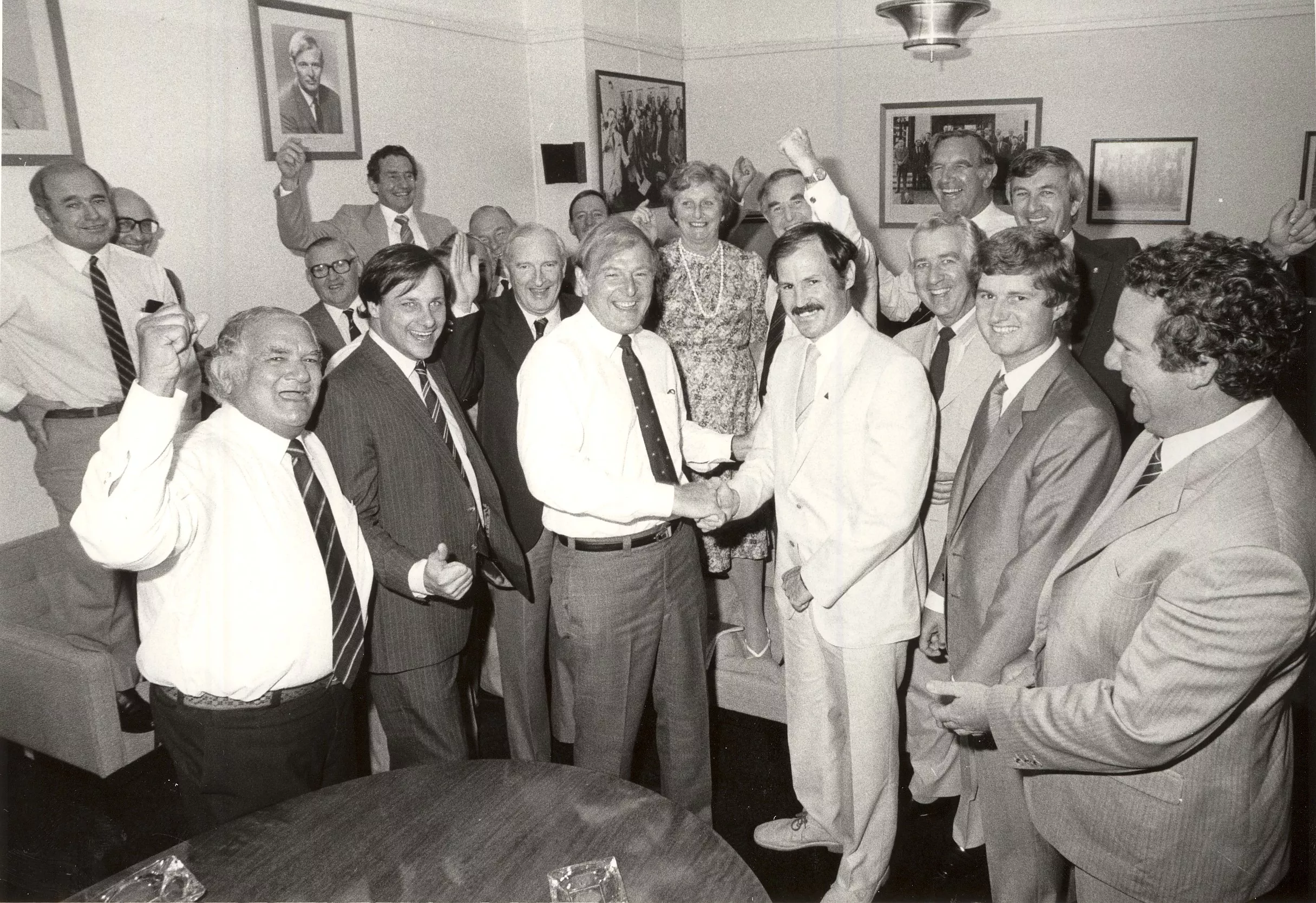 This black and white photograph captures the jubilation in the National Party Room on the first day back after the March 1983 federal election. Leader Doug Anthony shakes the hand of Noel Hicks, member for Riverina. Some of Noel’s colleagues have their hands raised in fist pumps and Flo Bjelke-Petersen is standing on a sofa with a big grin on her face. In the background are portraits of leaders and formal and informal photographs of the party.