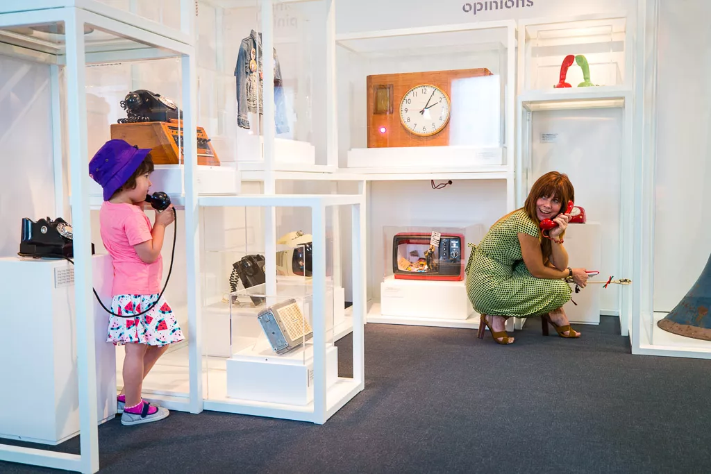 A child speaks on a phone talking to their mother surrounded by object cases in a museum with a clock, a radio and a TV. 