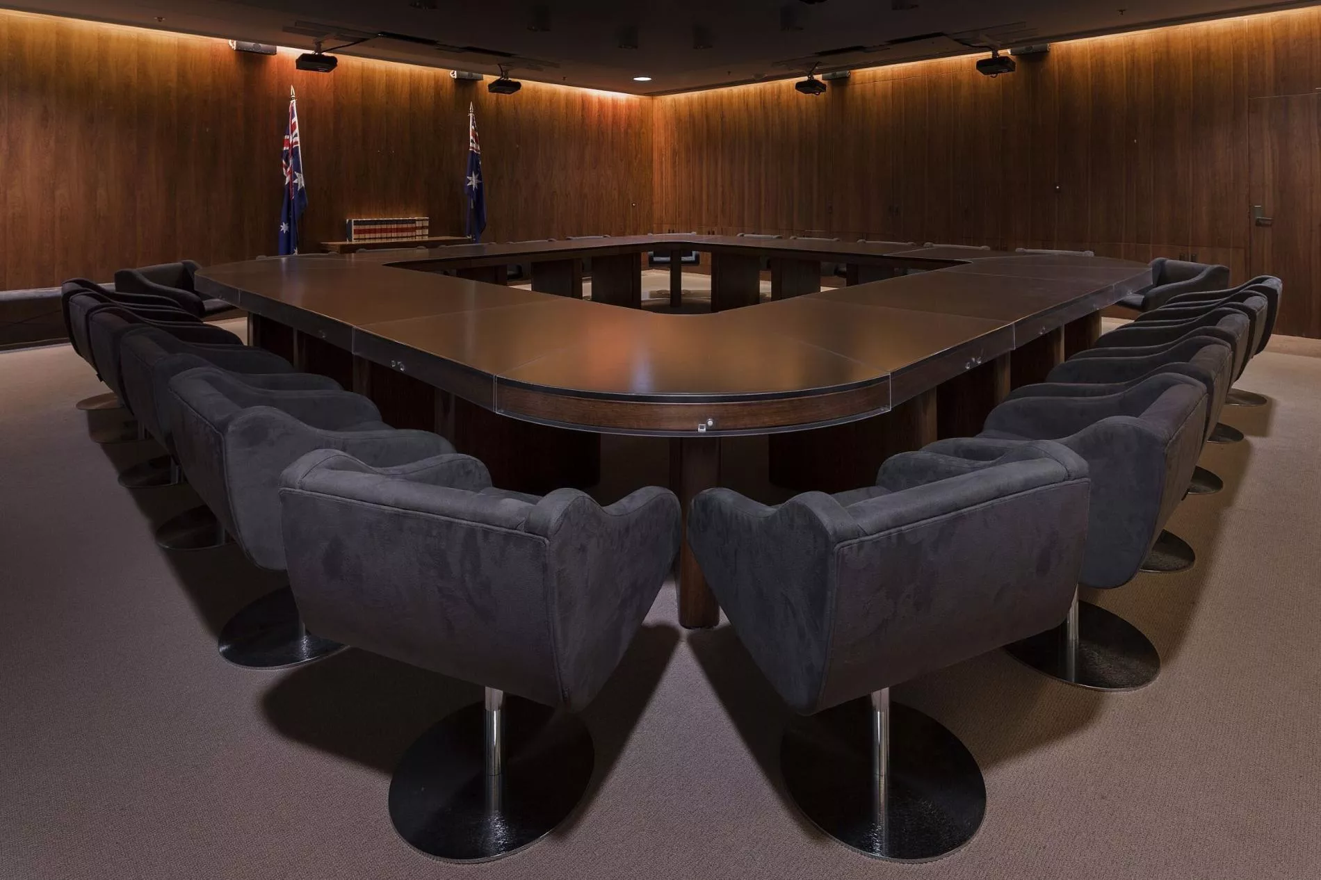 A dark room with heavy curtins and a large, heavy oval shaped table with a hole in the middle surrounded by large grey chairs. 