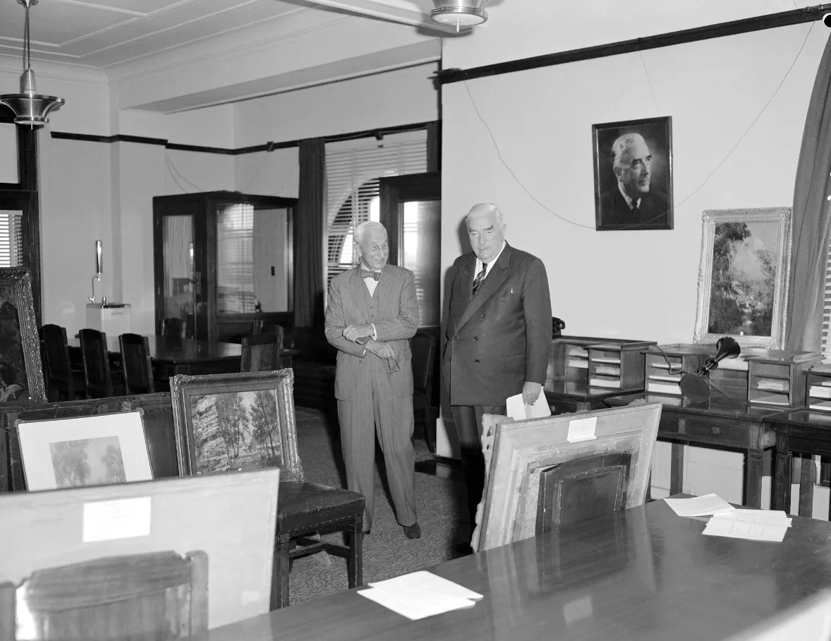 This black and white photograph shows two suited men standing side by side in the Government Party Room in 1962. On the left is Sydney businessman and philanthropist SH Ervin with a jaunty bow tie, on the right a more conservatively dressed Prime Minister Bob Menzies. They are surrounded by Australian paintings which are leaning against chairs, tables and walls. The two men are viewing the paintings that Ervin was donating to the National Collection. In the background are meeting tables and telephone booths