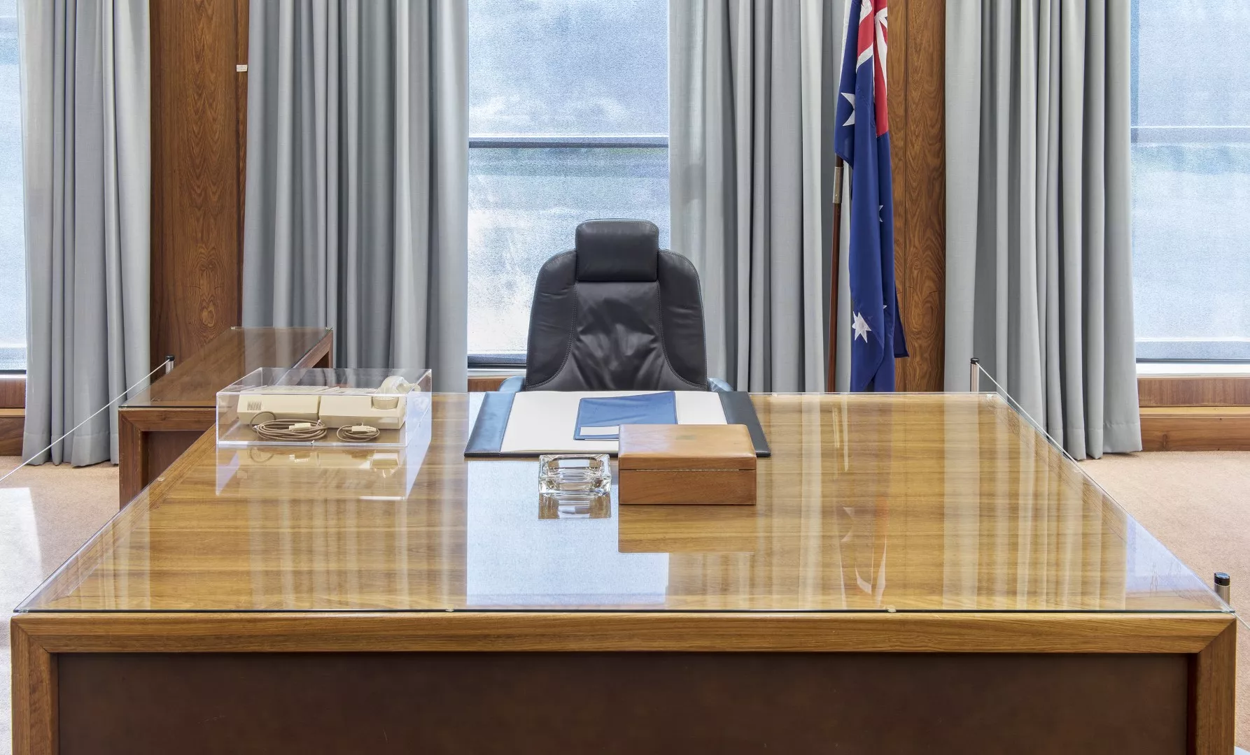 A front view of the Prime Minister's desk with a chair behind it. On the desk there is a 1980s telephone, blotter, glass ashtray and timber box.