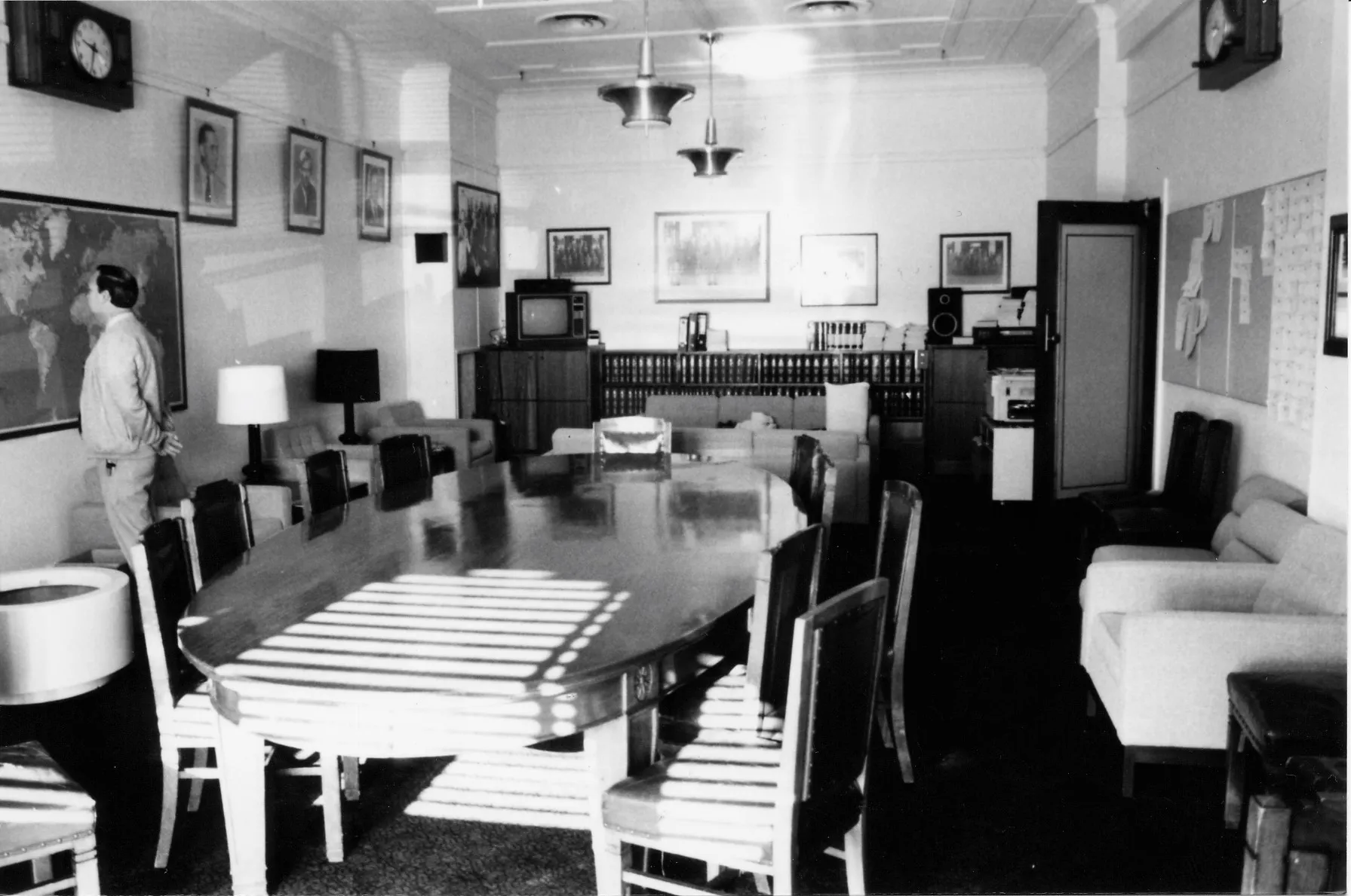A large oval meeting table and chairs dominate the centre of the room. Easy chairs with side tables and lamps are arrayed along the sides and two sofas are arranged in a seating area towards the back of the room. A sideboard with books, a television and sound system stretches along the back wall. There is a framed map on one wall and a notice board on the opposite. Portraits of leaders and formal and informal photographs of the party crowd the walls