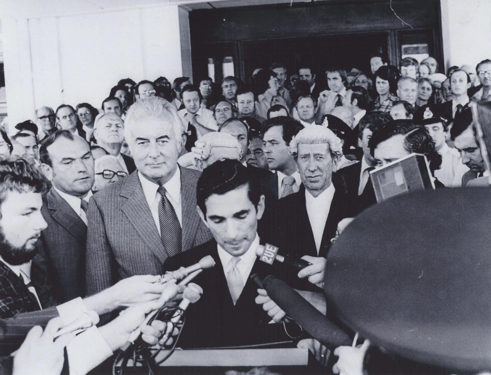 David Smith reads the dismissal proclamation outside Parliament House in 1975. Gough Whitlam stands behind him amongst a huge crowd. Reporters hold microphones.