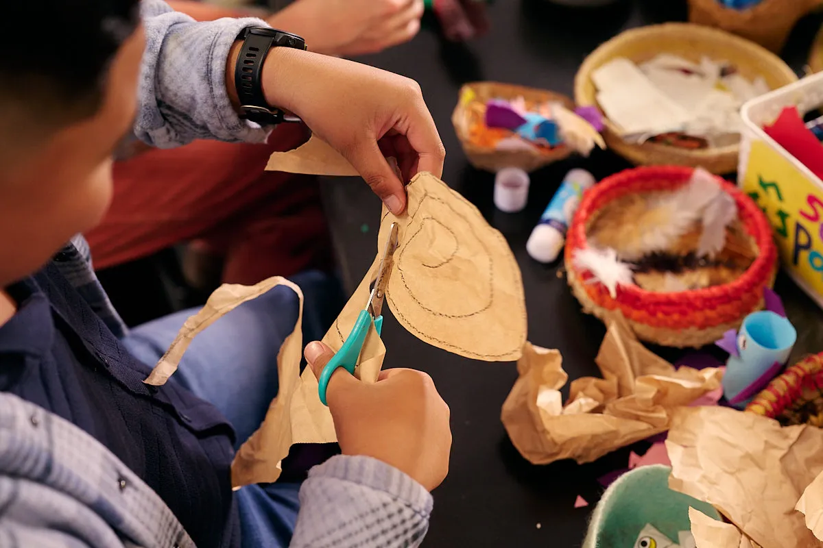 A kid cutting up some craft paper with scissors above a busy crafting table filled with items like glue, feathers, and paper.